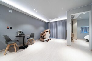 Quarto moderno com poltrona de massagem, uma mesinha redonda com cadeiras e uma grande janela com cortinas.