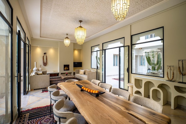 A spacious living room with a long wooden table, comfortable seating, and large windows offering a view of the outdoors. - Riad Malaika Marrakech (Marrakech)