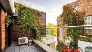A balcony with a wooden floor, a glass railing, and a view of a brick building with ivy. - The Primrose Hill Elegant House (Greater London)