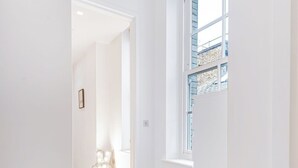 A hallway with wooden flooring and white walls, leading to a room with a window and a door.