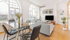 A modern living room with a glass dining table, grey sofa, and black leather chairs. A large TV is mounted on the wall, and there are framed pictures above it.