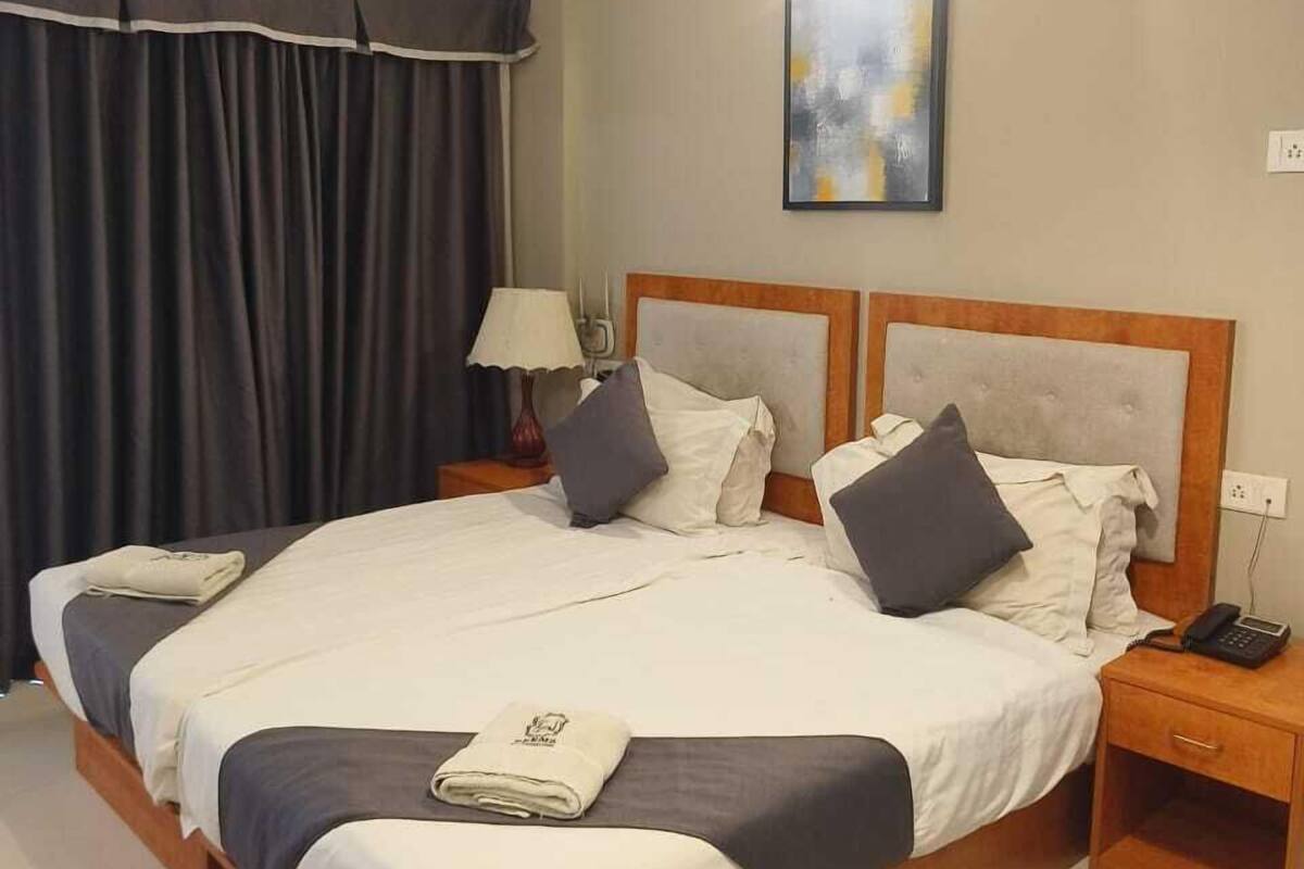 A hotel room with a large bed, a ceiling fan, an air conditioning unit, a bedside table with a lamp, and a painting on the wall.