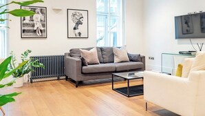 Una sala moderna con un sofá gris, un sillón blanco, una mesa de centro y un televisor. Hay cuadros enmarcados en la pared y un radiador abajo.