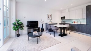A modern living room with a flat-screen TV, a dining area, and a kitchen with white cabinets and a dark backsplash. - All in One Gardened Flat (Greater London)