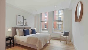 A modern bedroom with a large bed, a bedside table, a chair, and a window with a view of buildings.