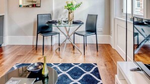 A modern living room with a glass dining table, two black chairs, a blue patterned rug, and framed pictures on the wall. - The Cosy Compact flat in Mayfair (Greater London)