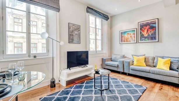 A modern living room with a flat-screen TV, a grey sofa with yellow cushions, a glass coffee table, and a patterned rug. - The Cosy Compact flat in Mayfair (Greater London)