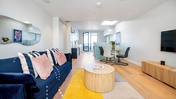 A modern living room with a blue sofa, a round wooden coffee table, and a dining area with chairs and a glass table. - Best Penthouse in Bloomsbury (Greater London)