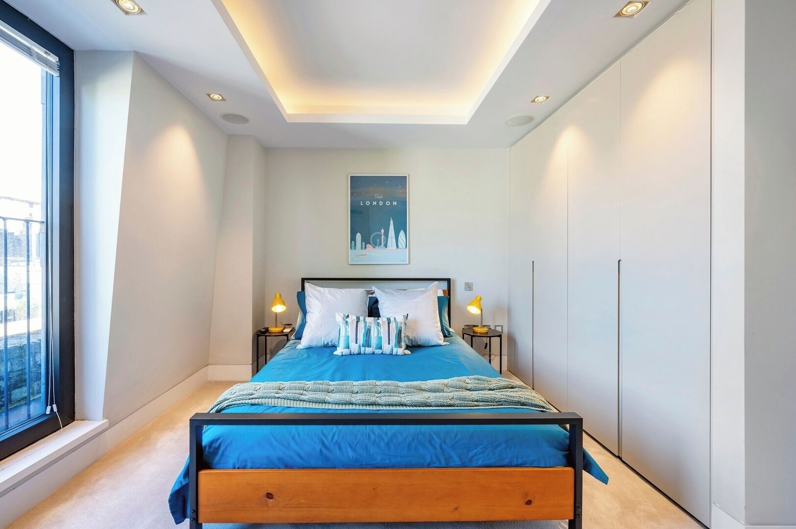 A modern bedroom with a large bed, two bedside tables, a skylight, and a cityscape poster on the wall.