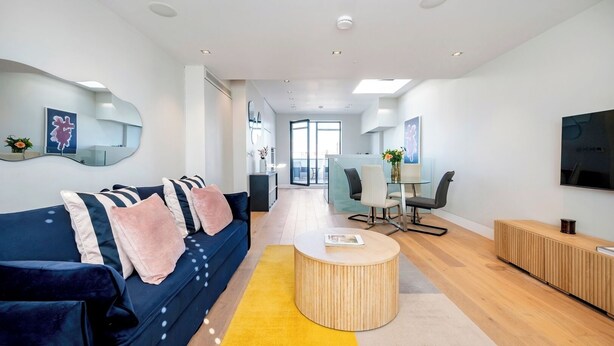 A modern living room with a blue sofa, a wooden coffee table, and a dining area with chairs and a glass table. - The Two Connected Penthouses (Greater London)