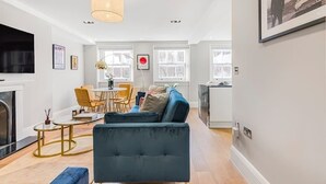 A modern living room with a blue sofa, a coffee table, and a fireplace. There is a dining area with a table and chairs, and a kitchen area with a counter and cabinets.
