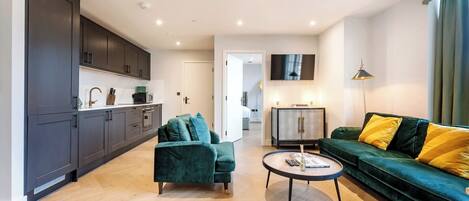 Ein modernes Wohnzimmer mit Sofa, Sessel und Couchtisch. Es gibt eine Küchenzeile mit Schränken und einer Mikrowelle. Ein Fernseher ist an der Wand montiert, und eine Lampe steht auf einem Beistelltisch.