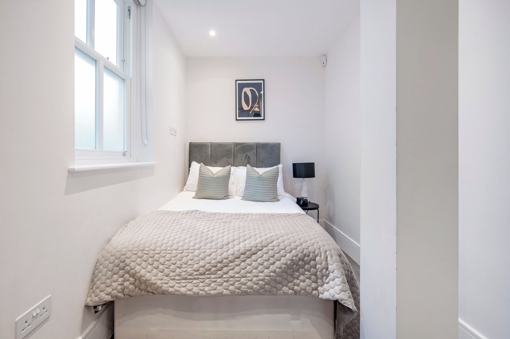 Een slaapkamer met een bed, nachtkastje, lamp, raam en een lijst met een afbeelding aan de muur.