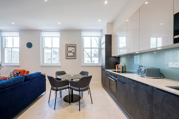 A modern kitchen with a dining area, featuring a blue sofa, a round table with chairs, and a large window providing natural light.
