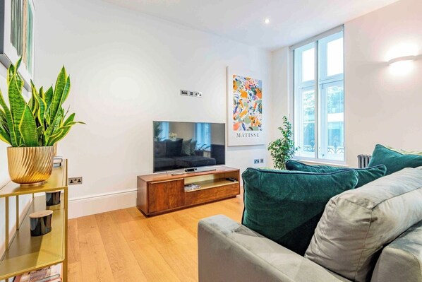 A modern living room with a flat-screen TV, a wooden cabinet, a green sofa, and a large window. - The 2 Connected Apartment in Covent Garden (Greater London)
