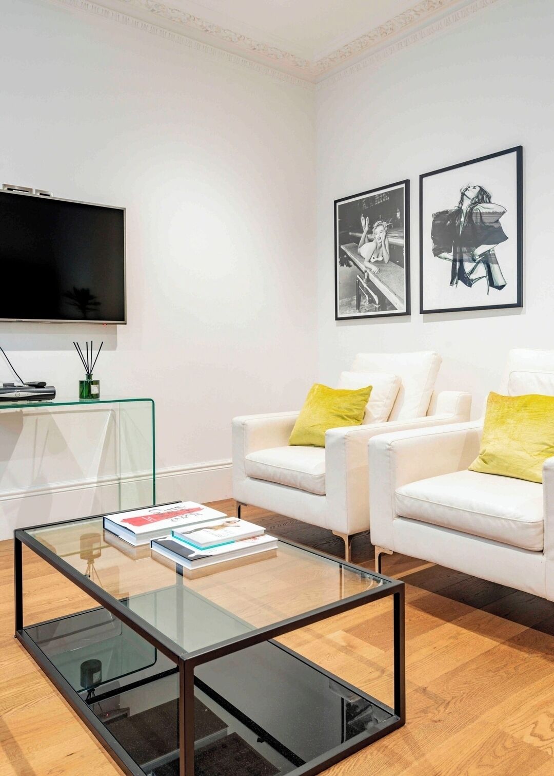 Ein modernes Wohnzimmer mit einem Glastisch, weißen Sofas mit gelben Kissen, einem Flachbildfernseher und zwei gerahmten Schwarz-Weiß-Fotos an der Wand.