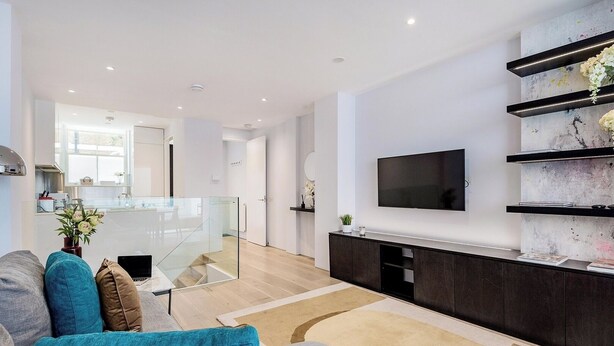 A modern living room with a sofa, a flat-screen TV mounted on the wall, and a glass staircase leading to the kitchen.