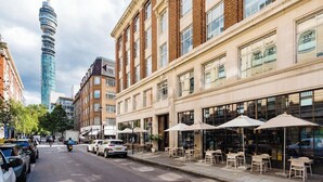 A street view with a row of buildings, including a tall tower, and outdoor seating areas with umbrellas.