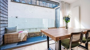 A modern dining area with a wooden table and chairs, a vase with flowers, and a view of a brick wall and window.