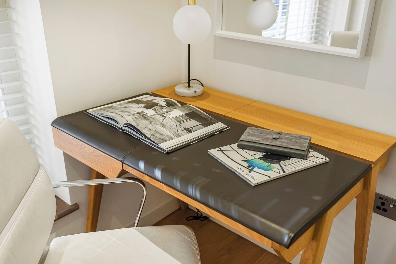 A modern room with a wooden desk, a black leather chair, a lamp, and a Louis Vuitton book.