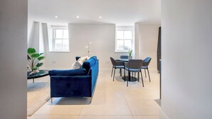 Un salon moderne avec un canapé bleu, une table à manger et des chaises, ainsi qu’une grande fenêtre laissant entrer la lumière naturelle.