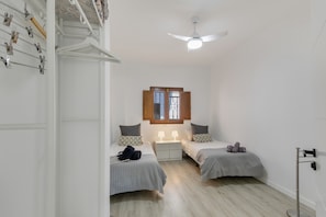Un dormitorio con dos camas, un ventilador de techo, una ventana con contraventanas, una mesita de noche y un estante montado en la pared.