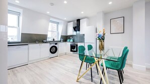 A modern kitchen with white cabinets, a washing machine, and a glass dining table with teal chairs. - Angel Brand New Penthouse (Greater London)