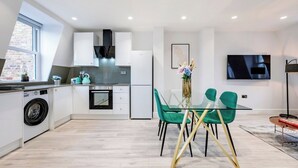 A modern kitchen with a built-in washing machine, a dining area with a glass table and teal chairs, and a flat-screen TV mounted on the wall. - Angel Brand New Penthouse (Greater London)