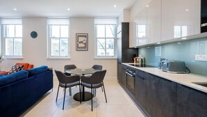 A modern kitchen with a dining area, featuring a round table with chairs, a blue sofa, and a large window providing natural light.