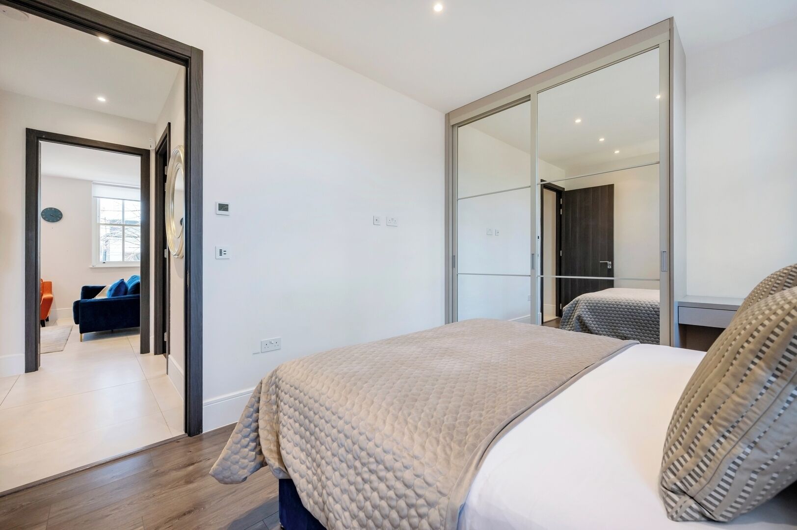 Une chambre moderne avec un grand lit, une armoire avec un miroir et une vue sur une autre pièce où se trouvent un canapé et une fenêtre.