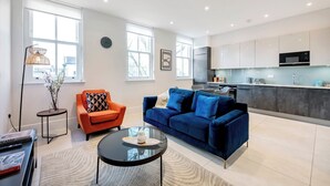 A modern living room with a blue sofa, orange armchair, and a glass coffee table. A kitchen with white cabinets and a black microwave is visible in the background.