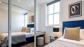 A modern bedroom with a large mirror, a bed with a blue headboard, a bedside table with a lamp, and artwork on the wall.