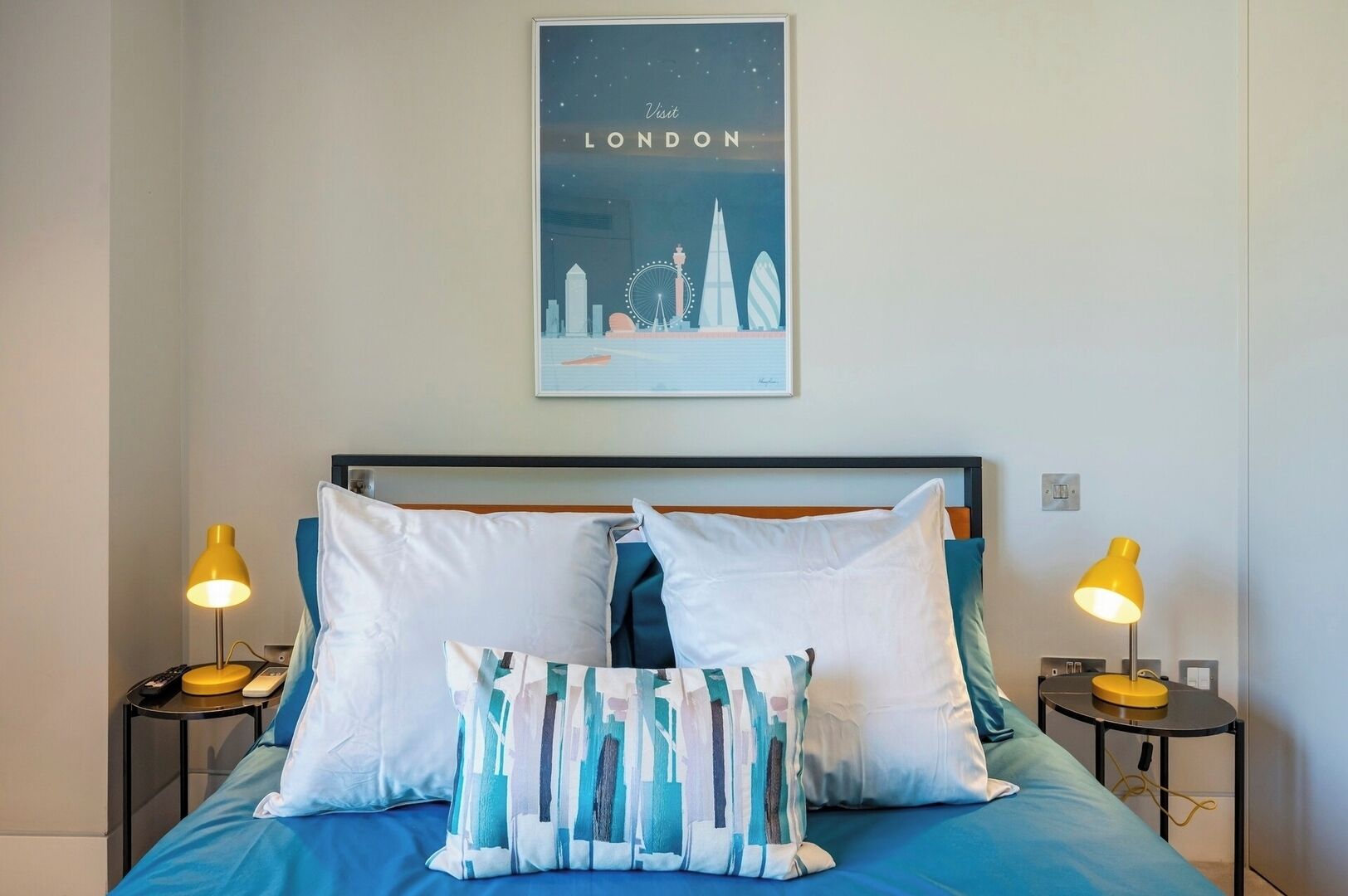 A hotel room with a bed, two bedside tables, and a poster of London landmarks on the wall.