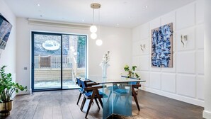 A modern dining area with a glass table, blue chairs, and a view of a brick building through glass doors. - Deluxe and Peace in Primrose Hill (Greater London)