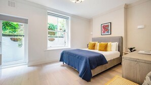 A bedroom with a bed, bedside table, and a window with a view of greenery. - Bloomsbury Gardened 2 bedFlat (Greater London)