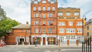 A multi-story red brick building with a corner entrance, a shop on the ground floor, and a gated driveway. - Bloomsbury Gardened 2 bedFlat (Greater London)