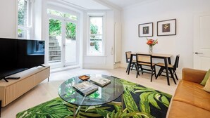 A modern living room with a flat-screen TV, a glass coffee table, a dining area with a wooden table and chairs, and large windows with a view of greenery. - Bloomsbury Gardened 2 bedFlat (Greater London)