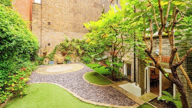 A courtyard with a brick wall, green grass, a gravel path, and trees. - Bloomsbury Gardened 2 bedFlat (Greater London)