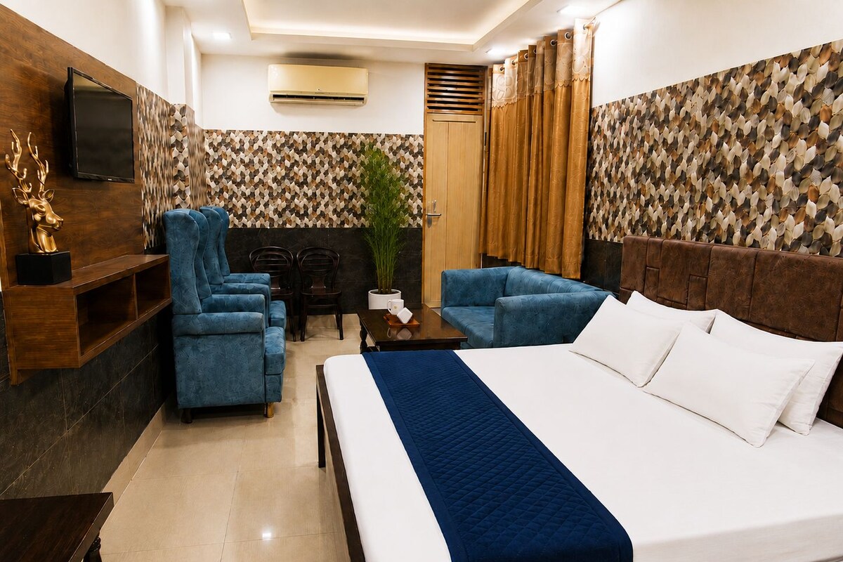 Habitación de hotel con cama, dos sillones azules, una mesita de madera, televisor montado en la pared y papel tapiz estampado.