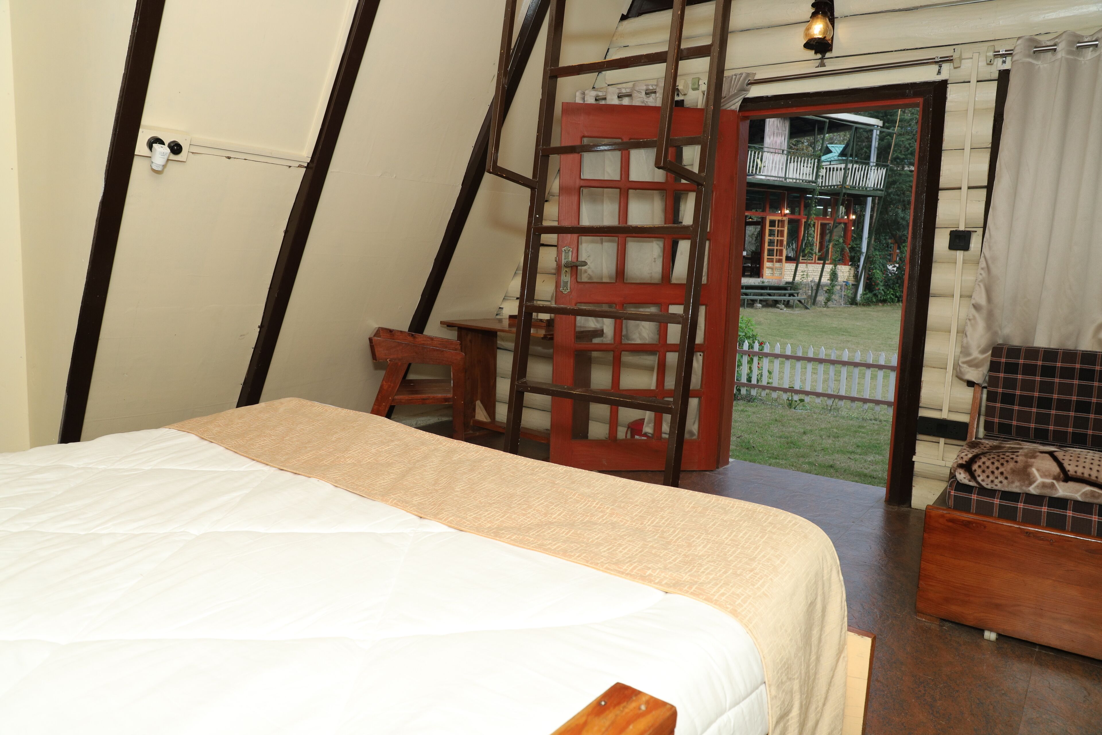 Una habitación con una cama, una silla de madera, una mesita y vistas a una casa a través de una puerta.