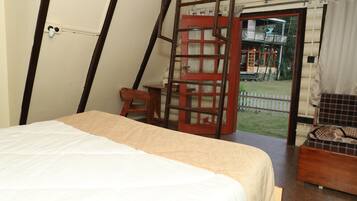 Una habitación con una cama, una silla de madera, una mesita y vistas a una casa a través de una puerta.