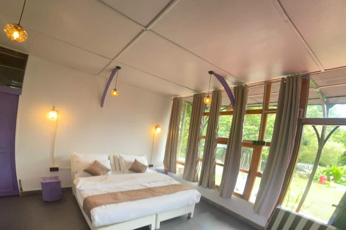 A bedroom with a bed, a small table, and a chair. There are windows with curtains, a lamp, and a view of greenery outside.