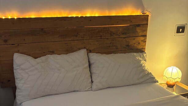 Ein ordentlich bezogenes Bett mit weißen Leinen und zwei Kissen, ein hölzernes Kopfteil, eine Nachttischlampe und ein kleiner Tisch mit einer Kerze.