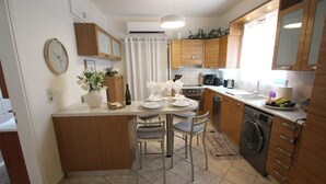 Een kleine keuken met houten kastjes, een inbouwoven, een wasmachine en een eethoek met stoelen en een tafel.