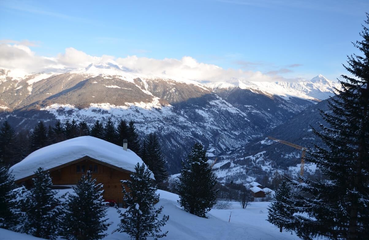 A wooden cabin with a snow-covered roof, surrounded by snow-covered mountains and pine trees.