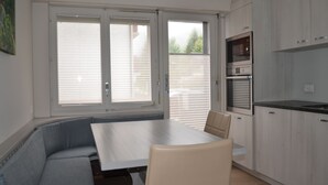 A modern kitchen with a dining area, featuring a built-in oven and microwave, a dining table with chairs, and large windows with blinds.
