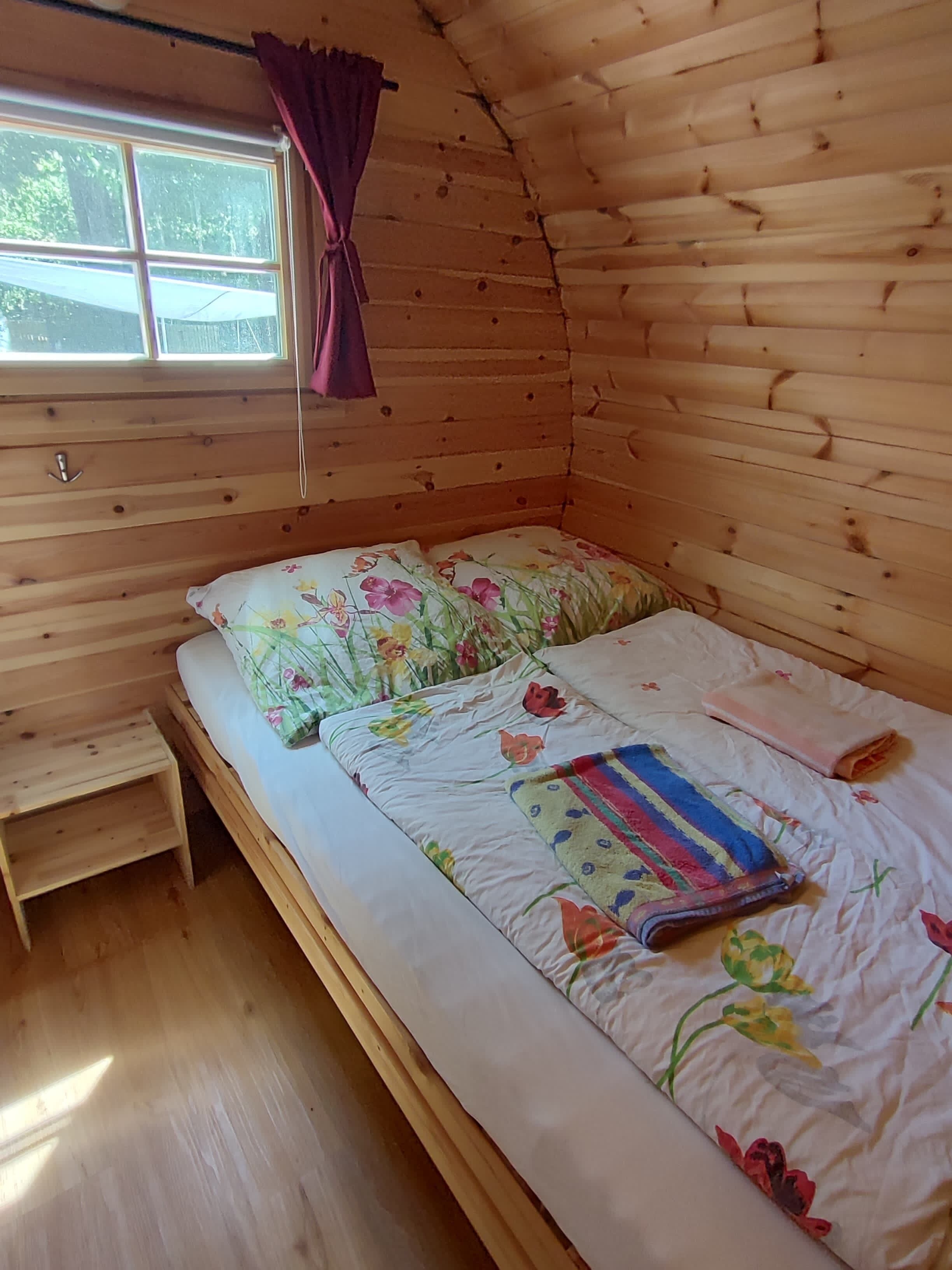 A wooden cabin room with a bed, a small wooden stool, and a window with a curtain.