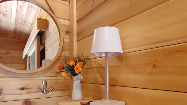 L'intérieur d'un chalet en bois, avec un miroir rond, une lampe blanche et un vase rempli de fleurs oranges, posé sur une étagère en bois.