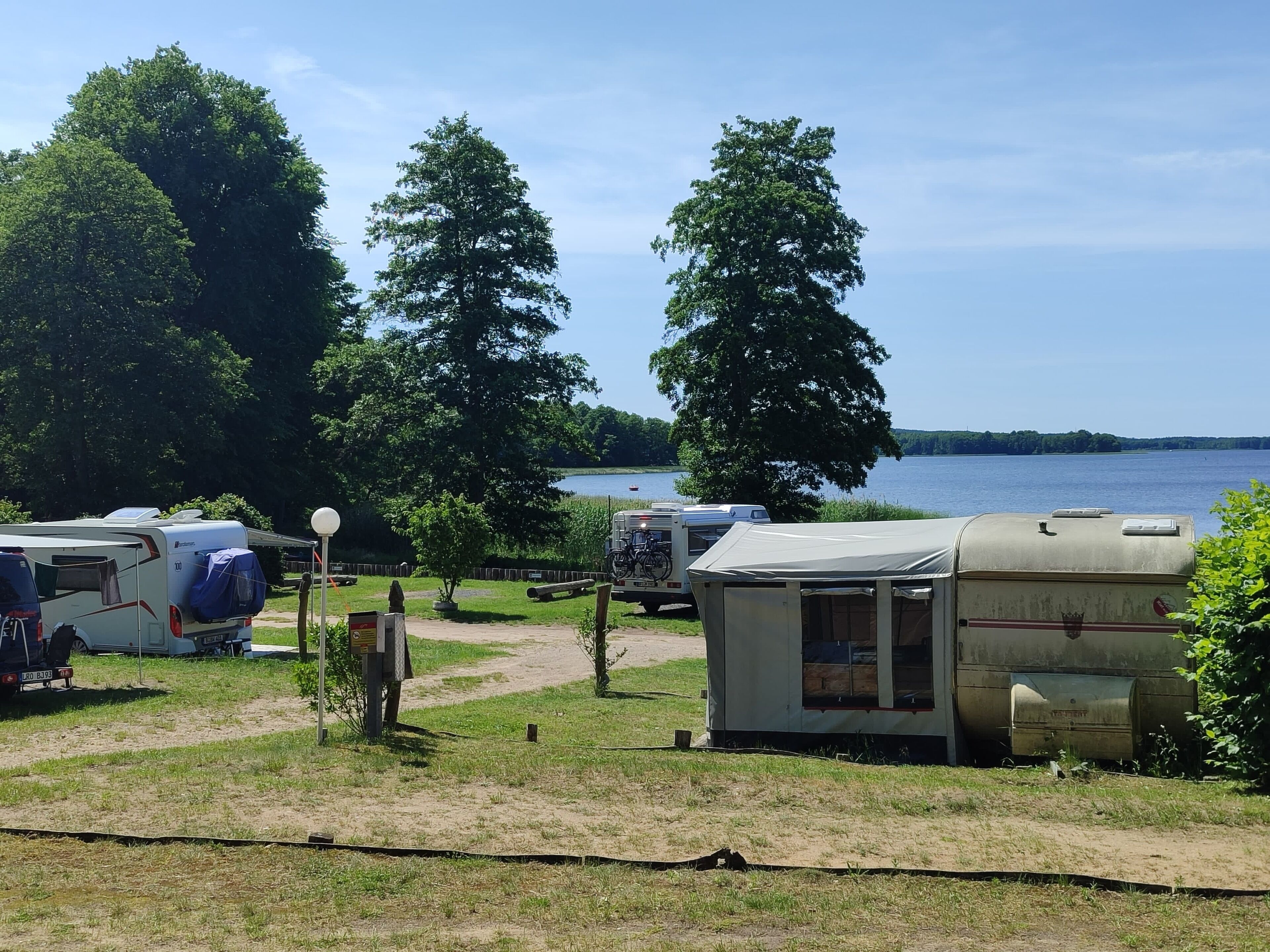 Un terrain de camping avec des camping-cars et une caravane, situé au bord d’un lac et entouré d’arbres.