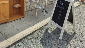 Un bâtiment à l’extérieur rouge et à la structure en bois, portant l’inscription « FRISCHE EIER » et contenant des collations et des boissons. Un panneau noir affiche la liste des articles proposés à la vente.